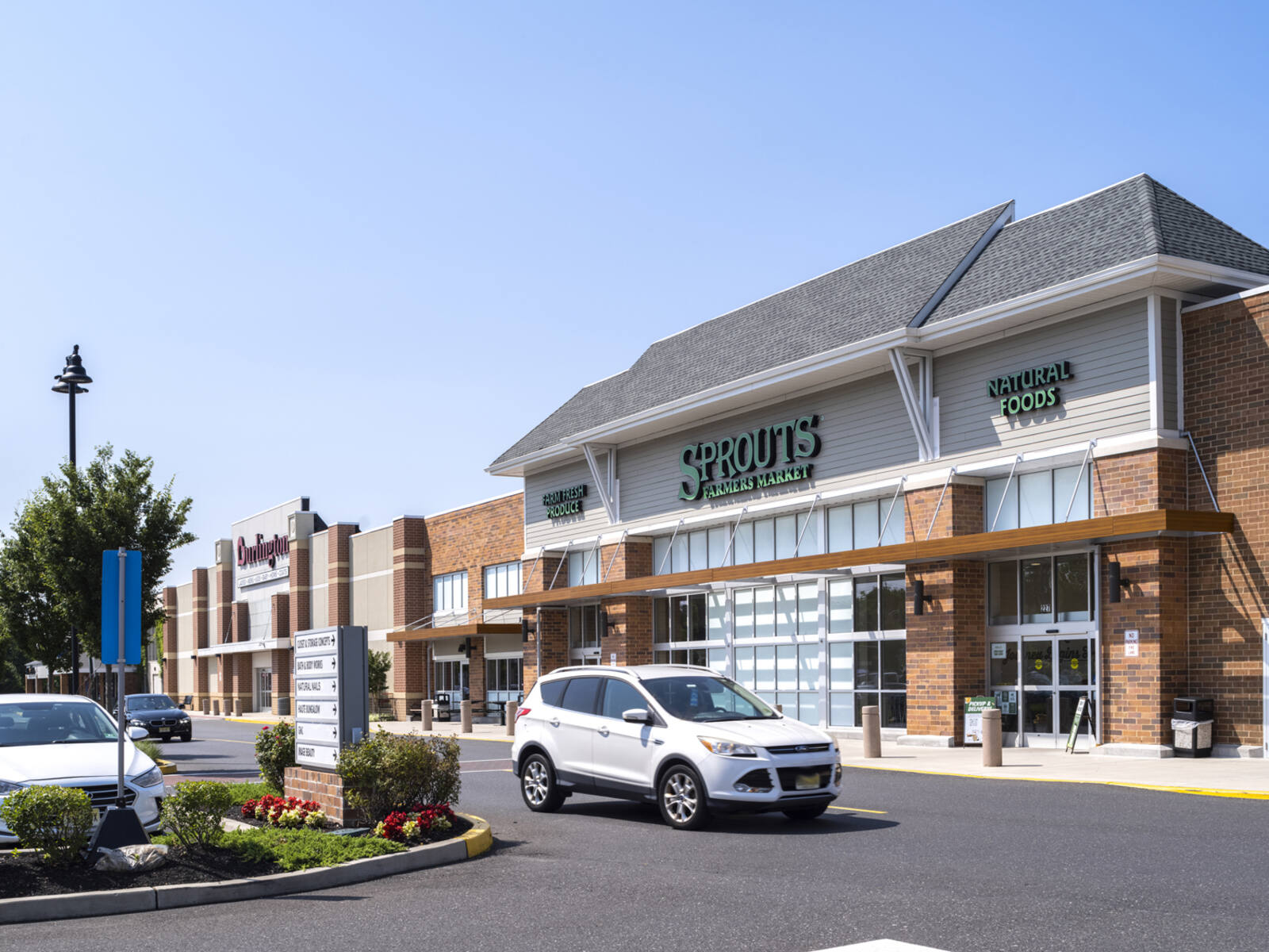White SUV stopped in front of Sprouts grocer in tree-lined parking lot.