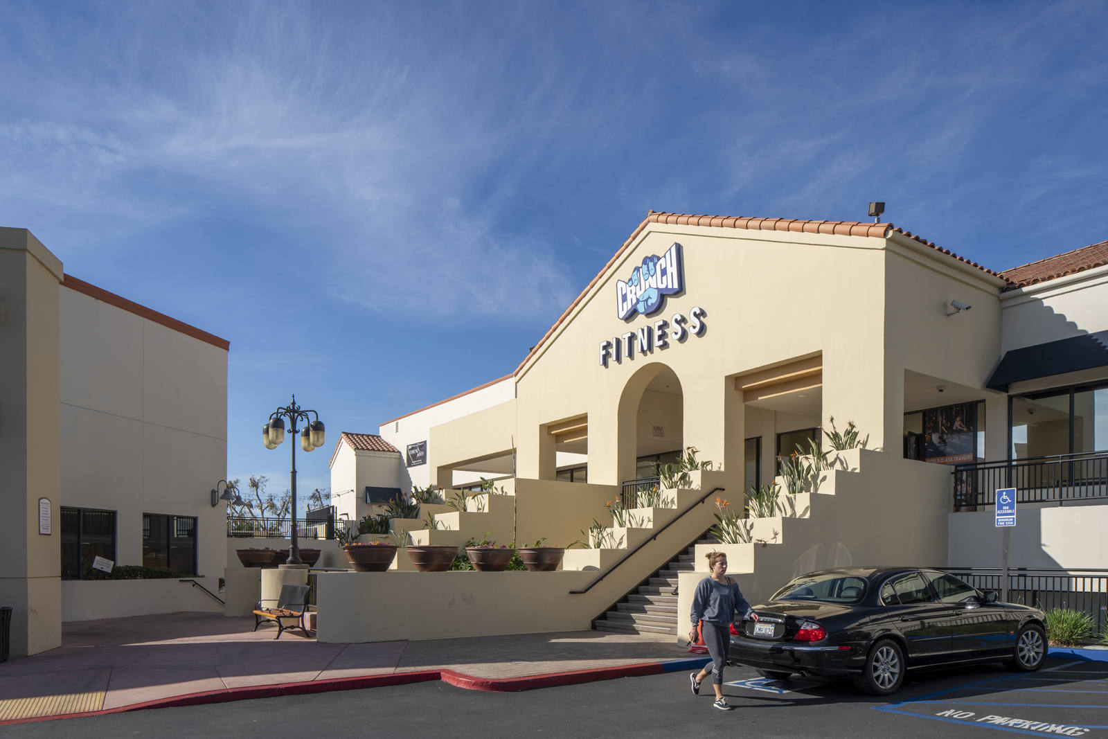 Person walking past black car in front of Crunch Fitness at outdoor shopping center