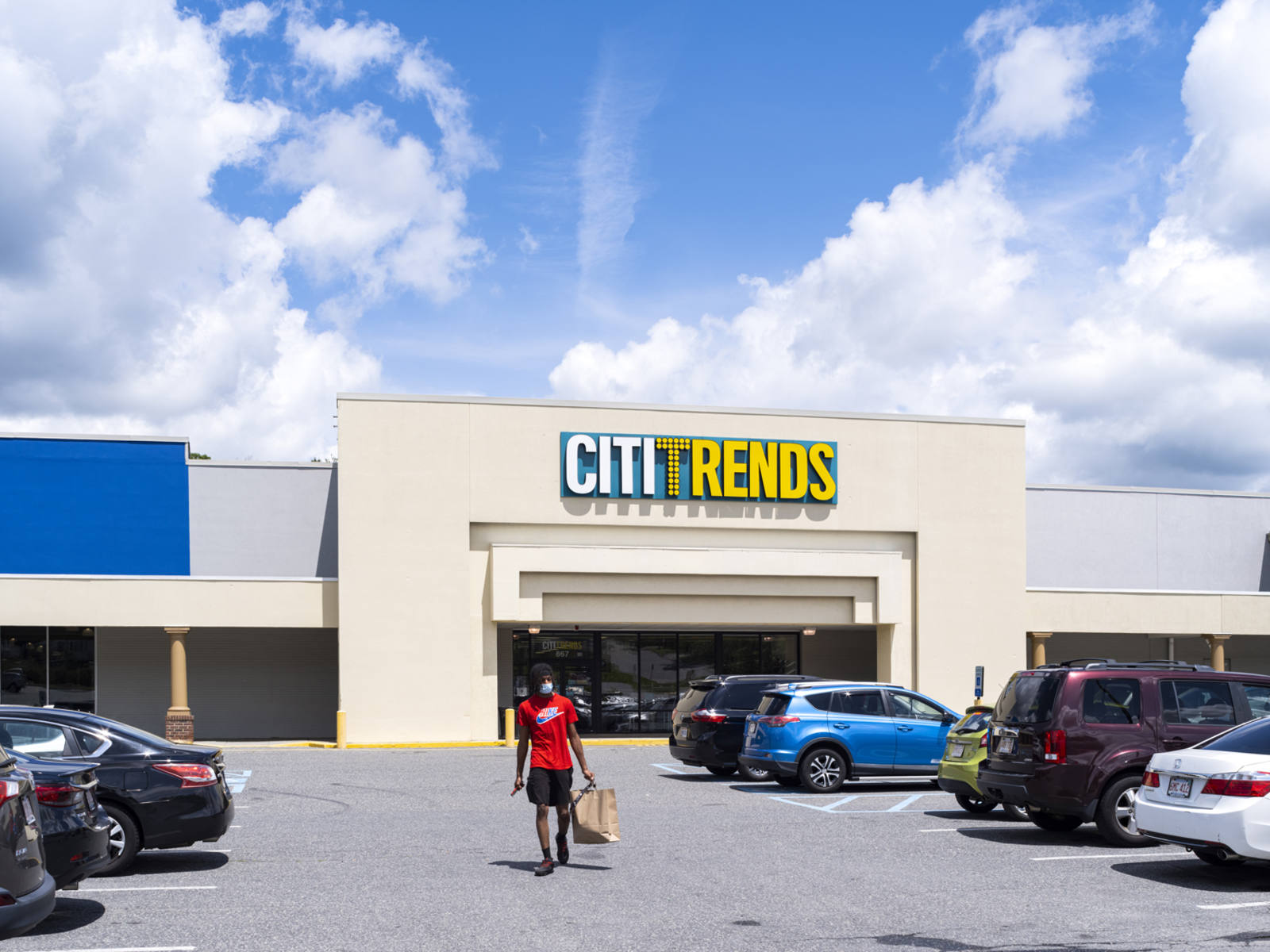 Person in red top carrying a grocery bag between rows of parked cars in front of Citi Trends