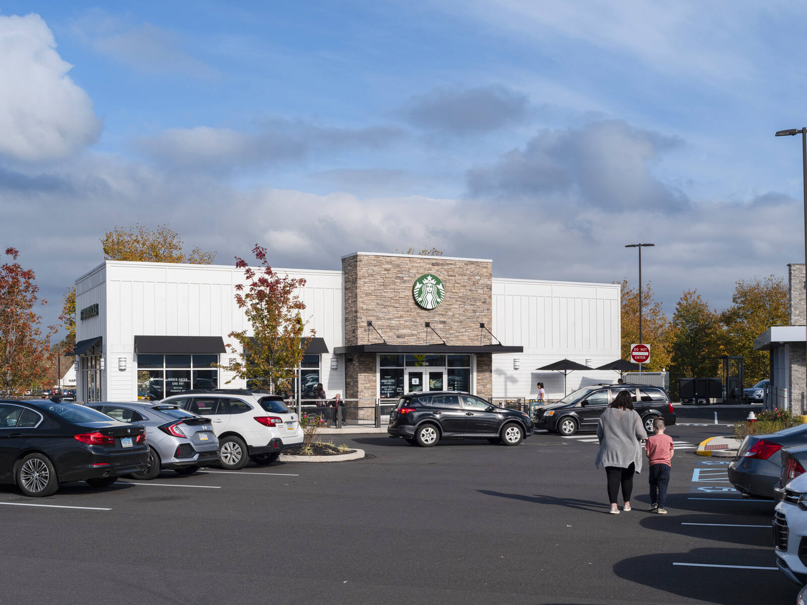 Adult and child walking through full parking lot in front of Starbucks with autumn fall trees throughout