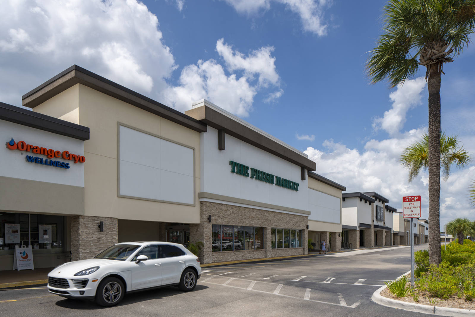 White car driving past front of The Fresh Market entrance with palm trees on the other side on a sunny day at the Park Shore Plaza shopping center