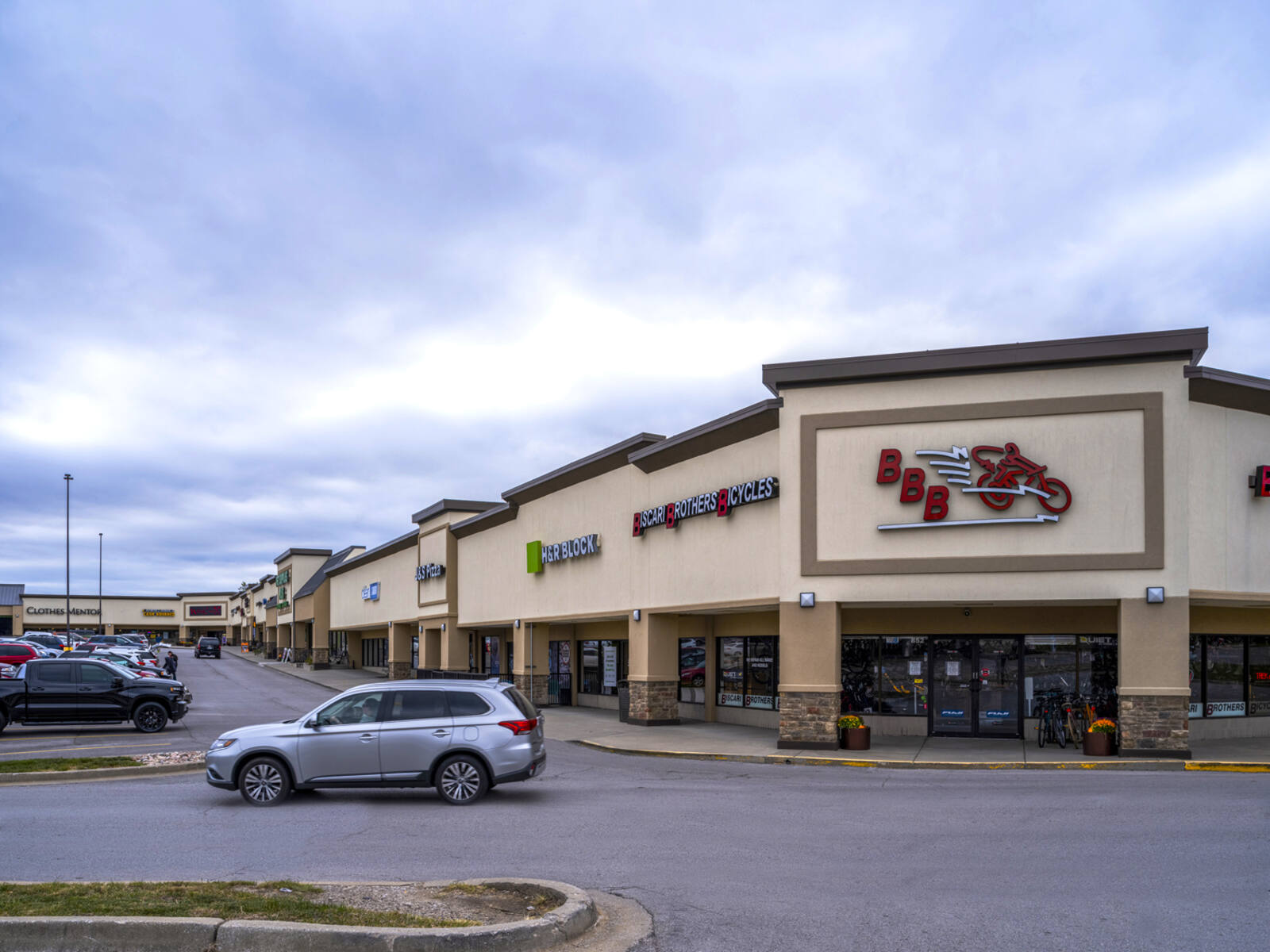 Biscari Brothers Bicycles and H&R Block with busy parking lot and grey SUV leaving lot.