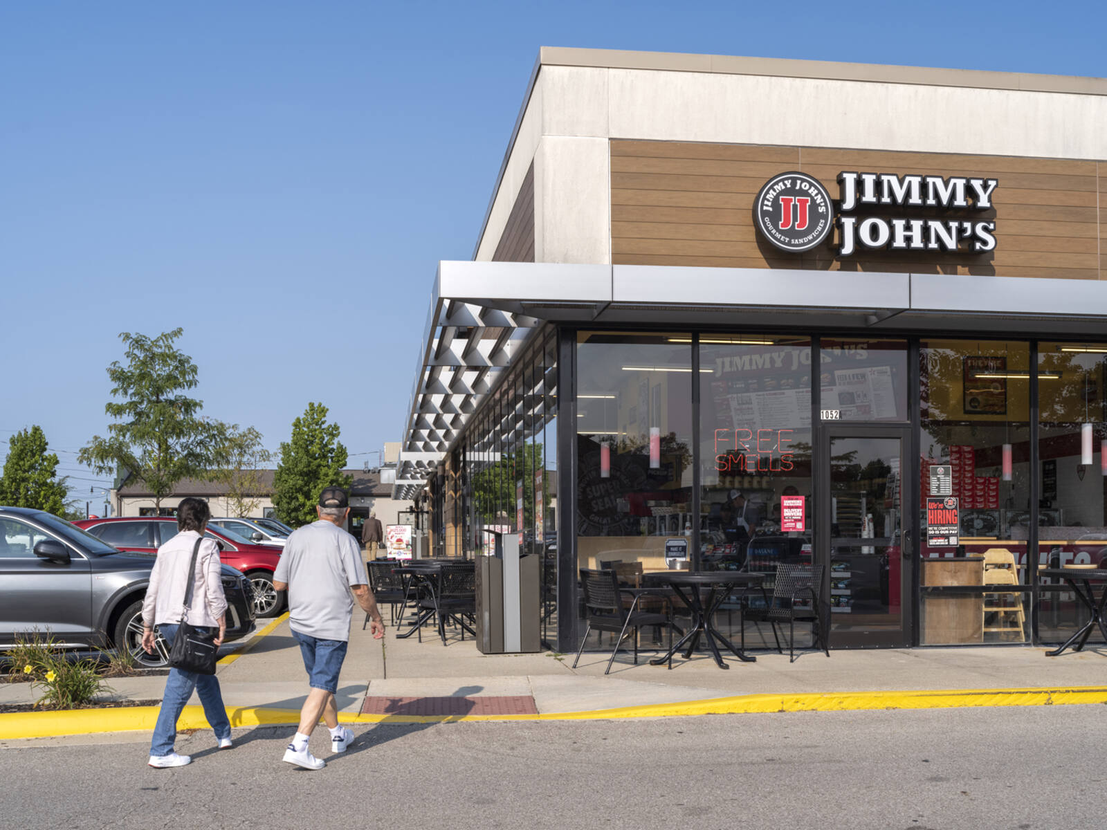 Two pedestrians approach Jimmy John's with outdoor seating and parking lot on left.