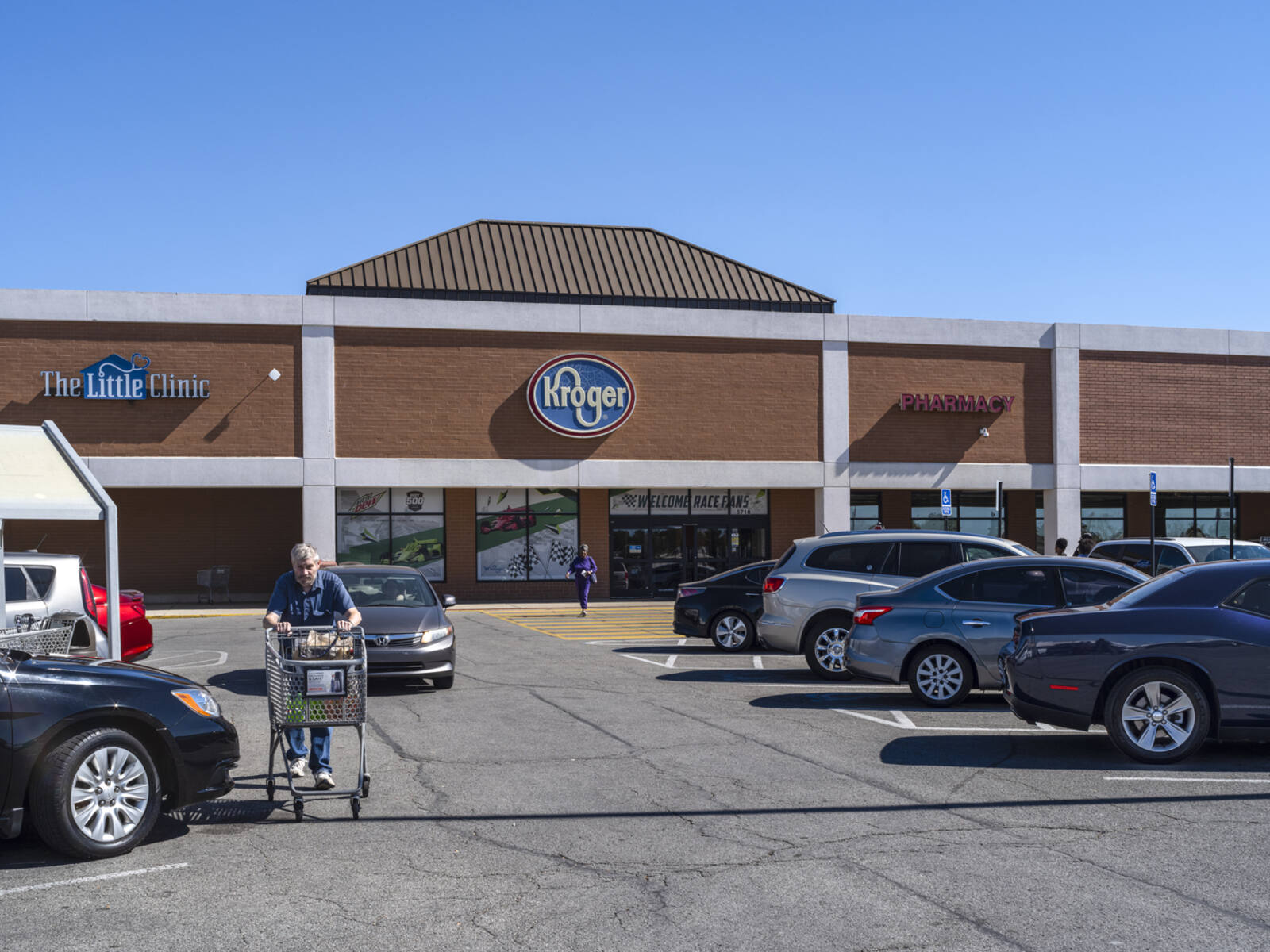 Man with shopping cart walking up busy parking lot aisle in front of Kroger.