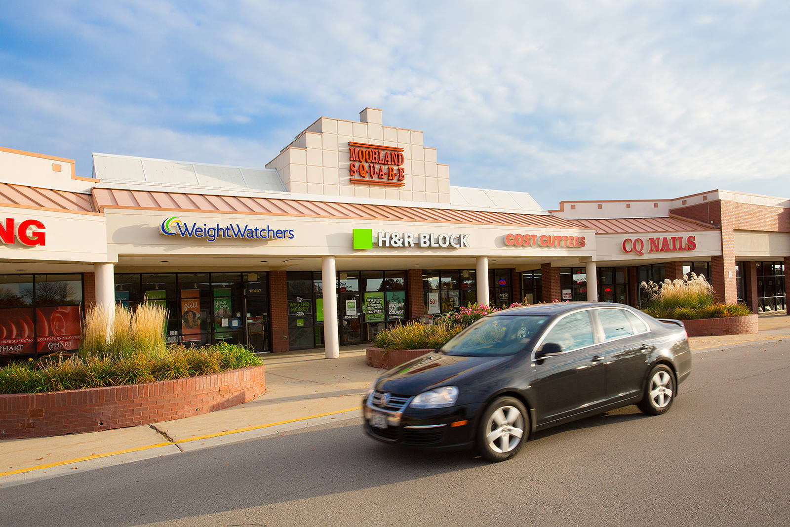 Black car passing HR Block and inline shops in New Berlin, WI.