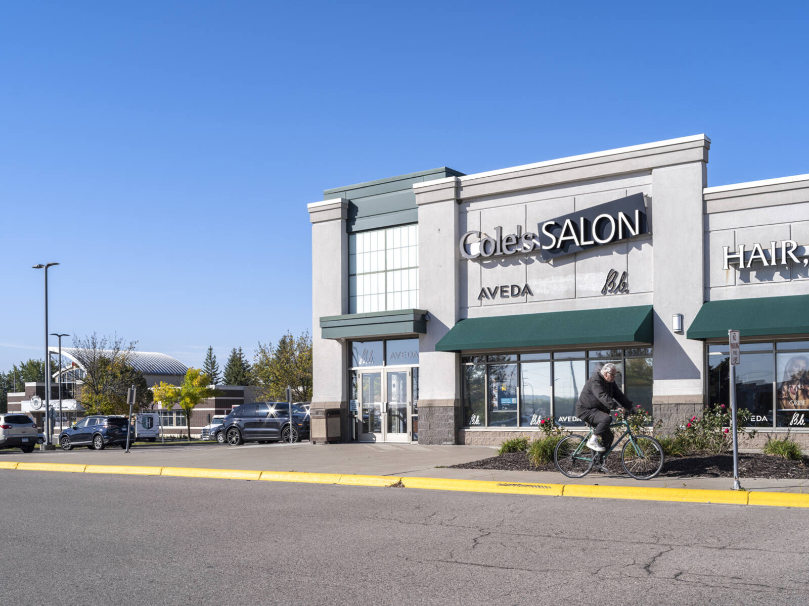 Man on bike passing Cole's Salon on sidewalk with parking lot in the background.