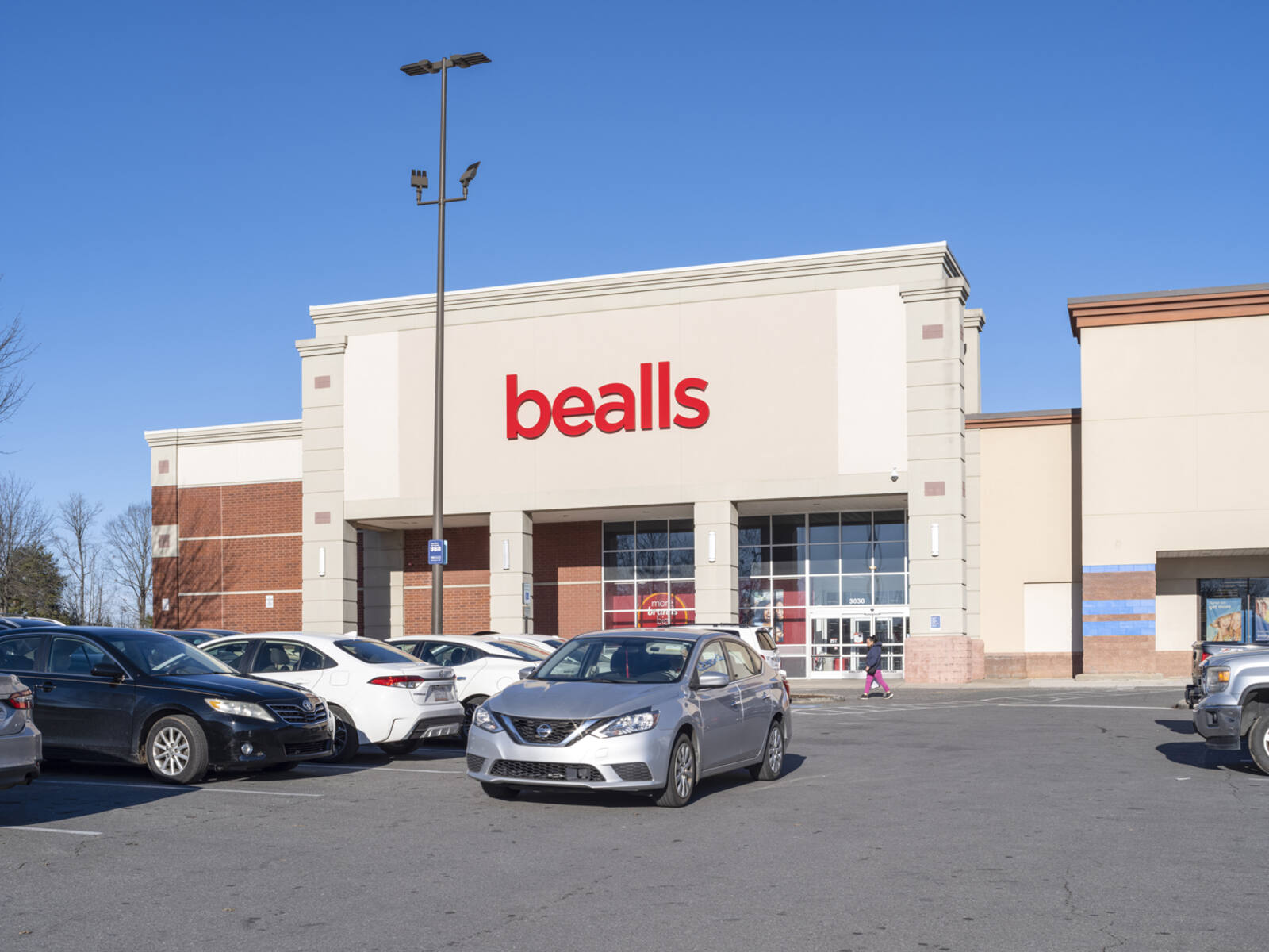 Silver car passing cars in front of bealls parking lot.