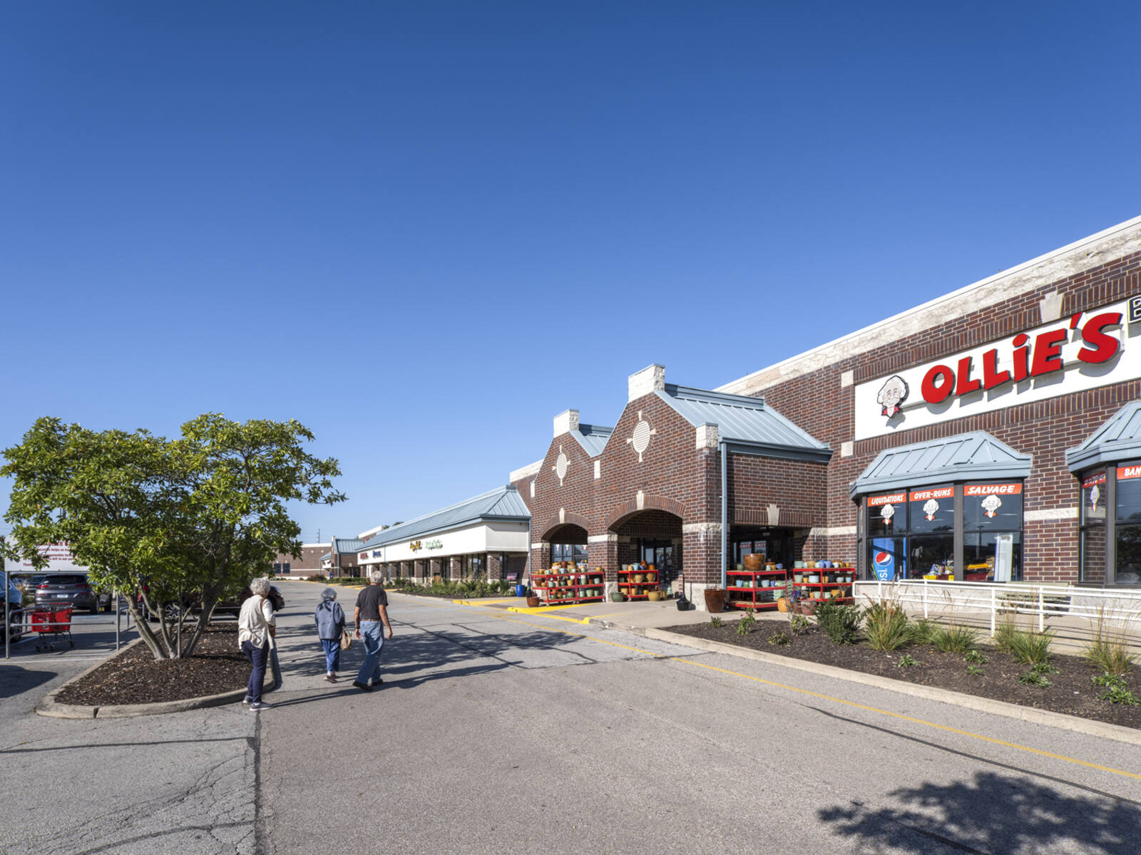 3 adults approach Ollie's with planters in front and trees by parking lot area.