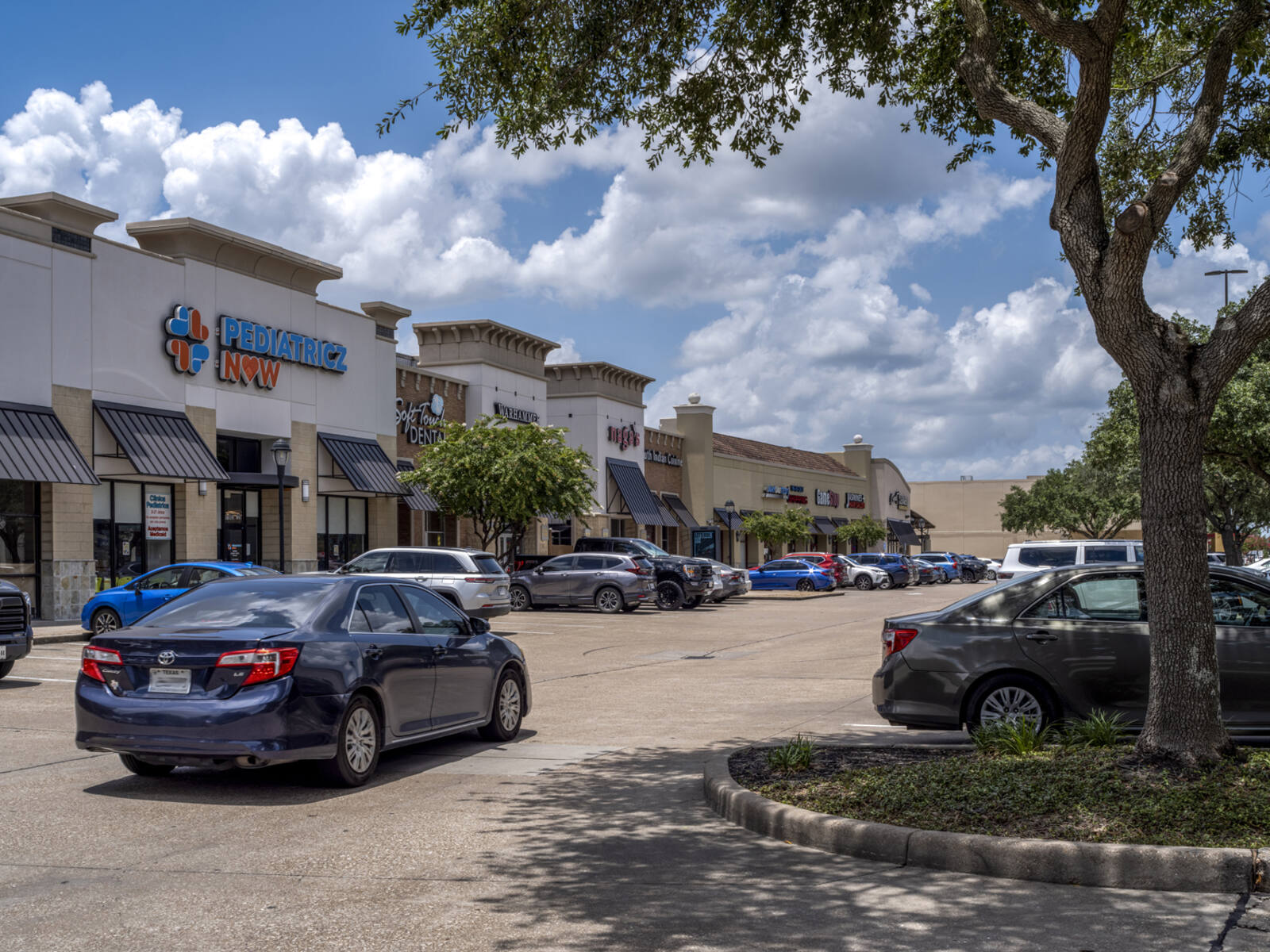 Blue car entering parking lot by Pediatricz Now and other stores.