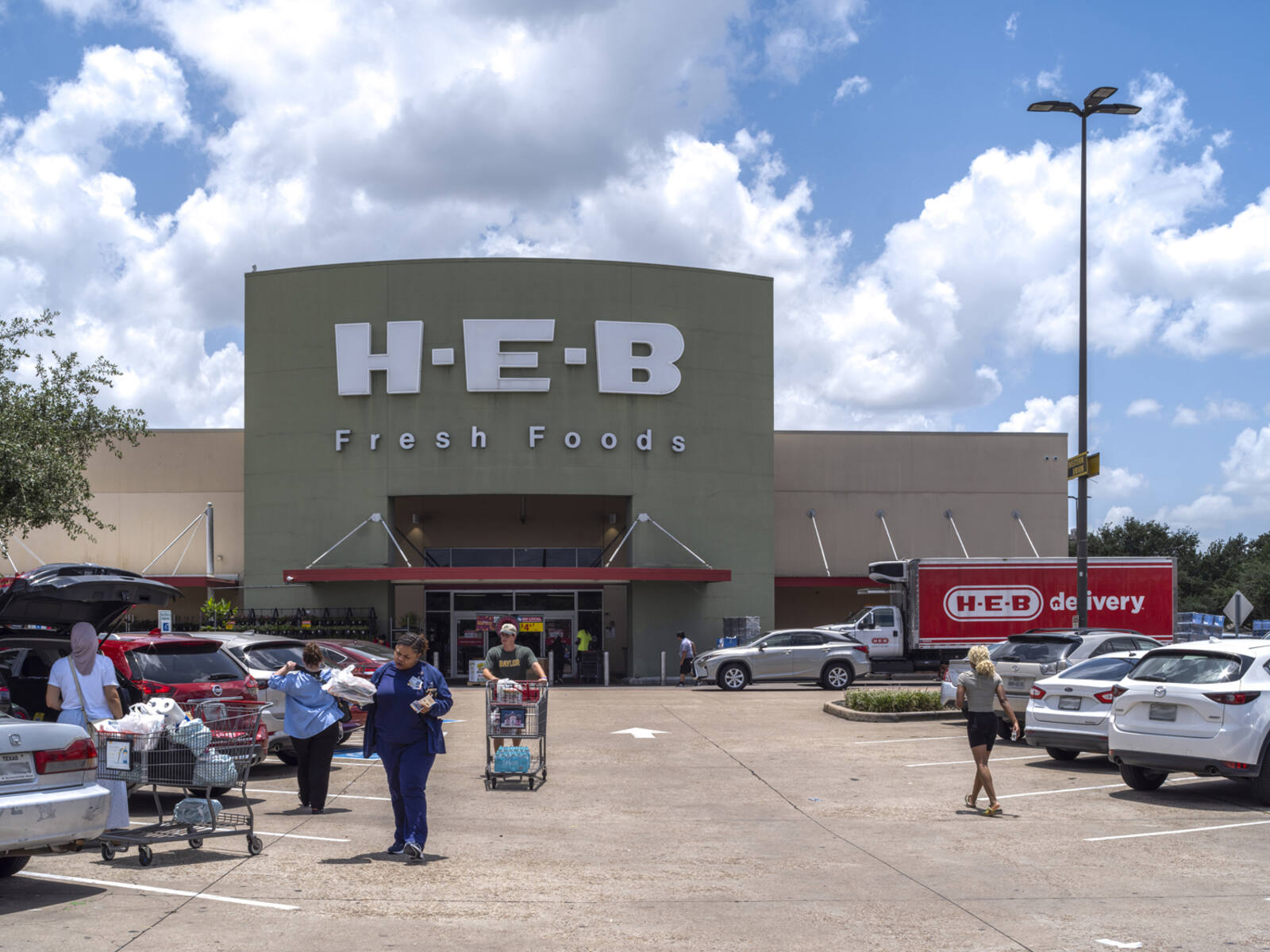 Shoppers with carts and bags in parking lot of busy HEB supermarket.