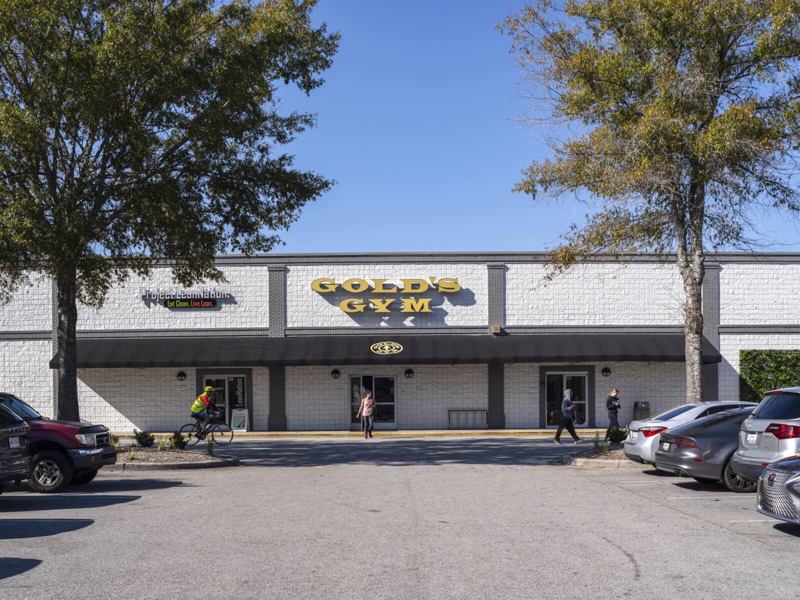 Bicyclist and pedestrians pass in front of Gold's Gym and parking lot.