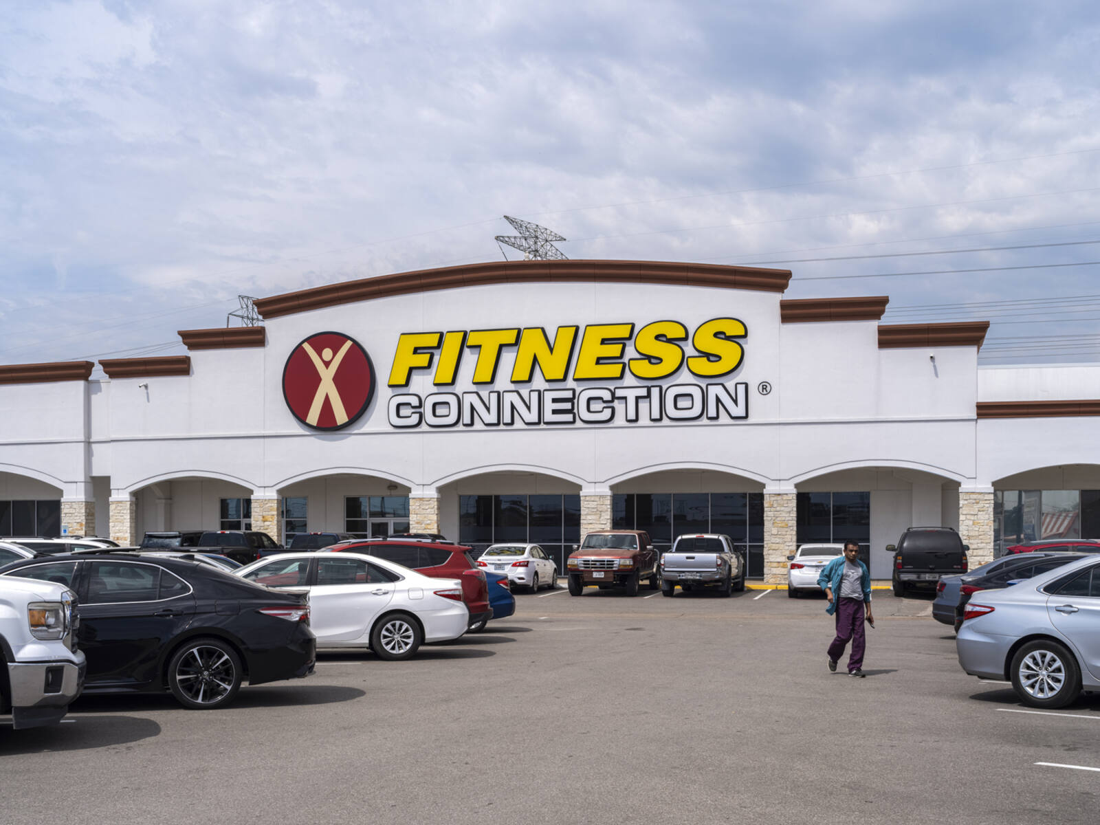 Busy parking lot at Fitness Connection with pedestrian walk down row of cars.