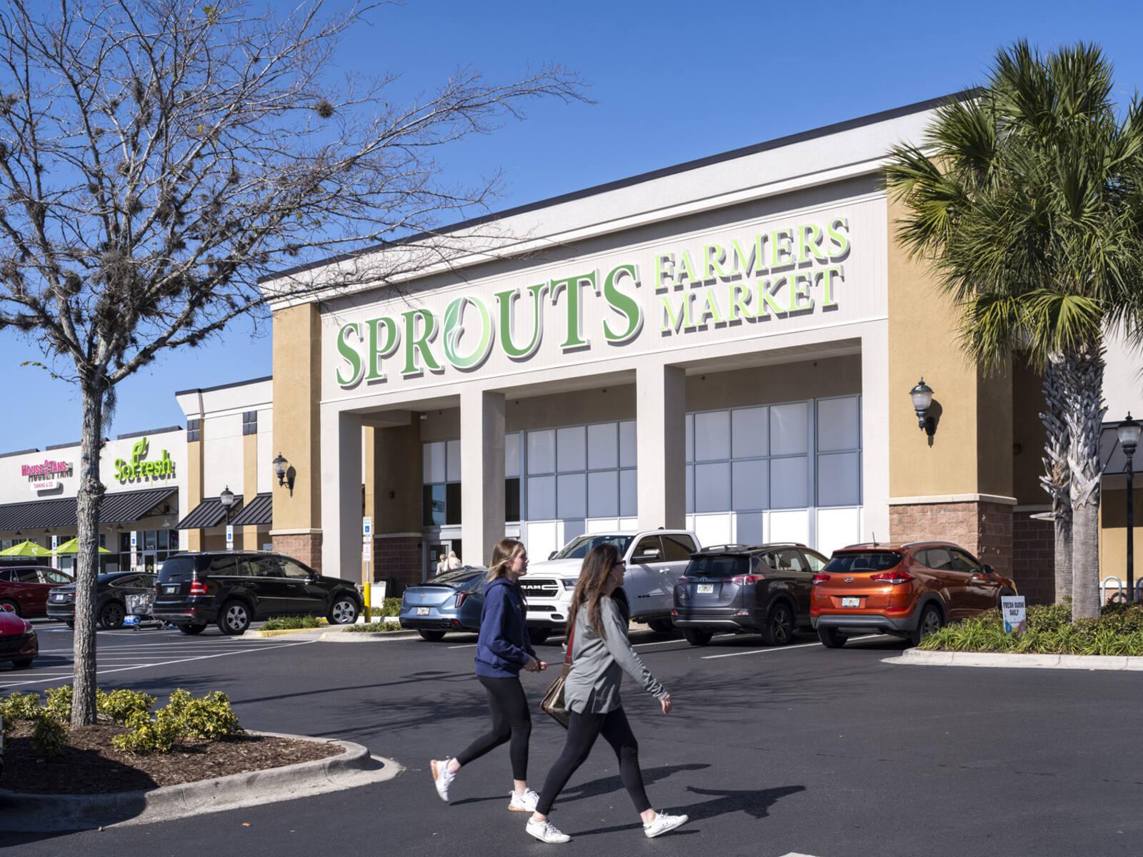 Two women approach Sprouts Farmers Market with cars and palm trees at entrance.