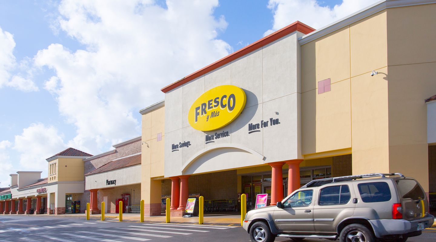 Fresco y Más at Shops at Palm Lakes in Miami, FL, featuring a yellow and cream façade with an arched entryway, bold oval signage, yellow safety bollards, a striped pedestrian crossing, and vehicles entering the front drive aisle beneath bright tropical daylight.