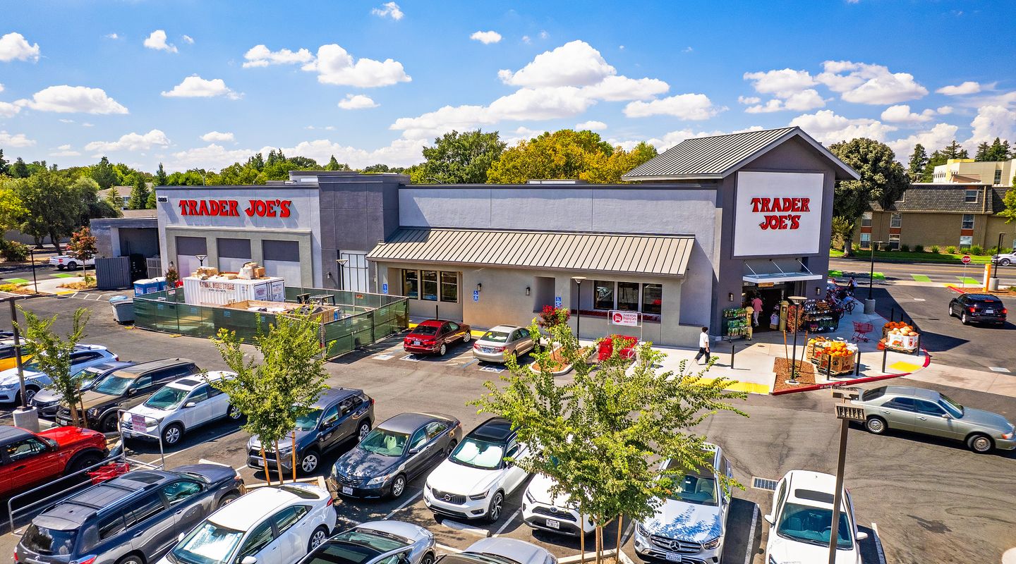 Trader Joe’s at The Davis Collection in Davis, CA, featuring a modern gray gabled façade with large white signage, outdoor seasonal produce displays, bike racks, landscaped walkways, and a wide pedestrian entry area under bright blue skies with scattered clouds.