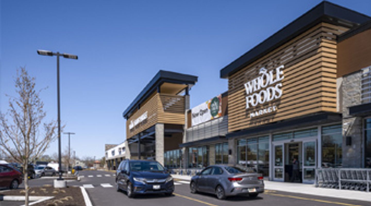 Two cars driving in front of Whole Foods Market storefront at Barn Plaza in Doylestown, PA, featuring modern architectural design, active customer traffic, and a newly paved parking area within the retail center.