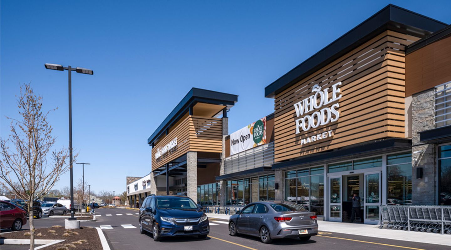 Two cars driving in front of Whole Foods Market storefront at Barn Plaza in Doylestown, PA, featuring modern architectural design, active customer traffic, and a newly paved parking area within the retail center.