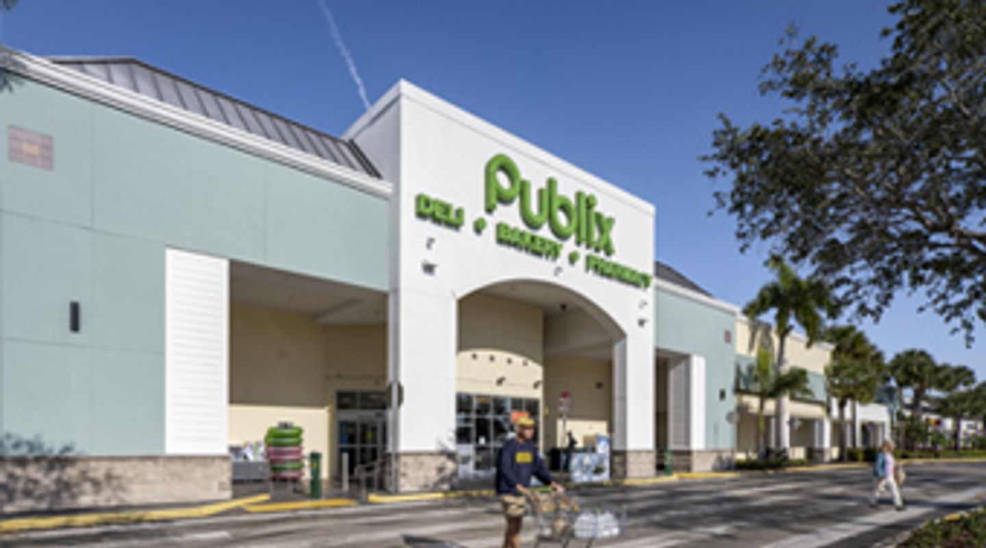 Publix supermarket at Freedom Square in Naples, FL, shown with a clean arched entrance, green signage, landscaped walkways, and shoppers moving through the parking area in front of the store.