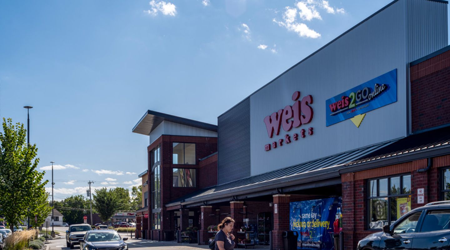 Weis Markets at Plymouth Square in Conshohocken, PA, shown with a contemporary red‑brick and gray metal façade, large glass entryway, covered walkway, prominent ‘weis2GO online’ signage, and vehicles circulating through the front drive aisle.