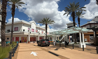 Daytime view of Pointe Orlando in Orlando, FL, featuring a wide pedestrian walkway lined with tall palm trees, a modern glass‑roofed shade structure, landscaped planters, and a two‑story retail and dining building with colorful window graphics and covered storefronts under a partly cloudy sky.