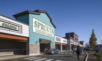 Sprouts Farmers Market at Roosevelt Mall in Philadelphia, PA, featuring a prominent green gabled façade with stone accents, large storefront windows, wide pedestrian crosswalk, landscaped walkways, and adjacent inline retailers under a clear blue sky.