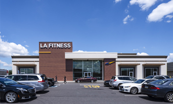 LA Fitness at Roosevelt Mall in Philadelphia, PA, featuring a modern exterior with red and white architectural finishes, large glass entrance, wide pedestrian walkway, and multiple vehicles parked in the foreground under a bright blue sky with scattered clouds.